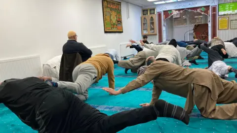 Cathy Minton/BBC A pilates class being held in a mosque in Bradford. The men participating are in comfortable, casual clothing and traditional dress.