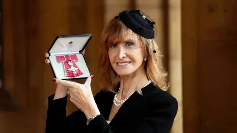 PA Media Jackie Bird, who has red hair, smiles at the camera as she shows off her MBE. It is in its case and attached toa  ribbon with a bow feature. Jackie is wearing a black jacket and black fascinator. She has a string of pearls around her neck.