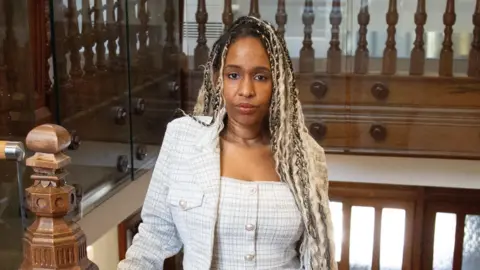 Kiki Marriott is stood inside the Parkland Place Rehabilitation Centre at the top of some wooden stairs. She has blonde braids and is wearing a white patterned suit. Kiki looks directly at the camera.