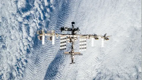 NASA The picture shows the entire International Space Station seen from directly above, floating over a textured blanket of white cloud. The station’s long central truss runs horizontally across the frame, with large rectangular solar panels fanning out to top and bottom like wings. The modules and structures along the spine form a dense, metallic knot at the centre, while smaller radiators and panels extend at angles near the ends. Below, Earth’s clouds appear as rippled, swirling patterns in pale blue and white, giving a strong sense of height and the station’s fragile isolation in orbit.