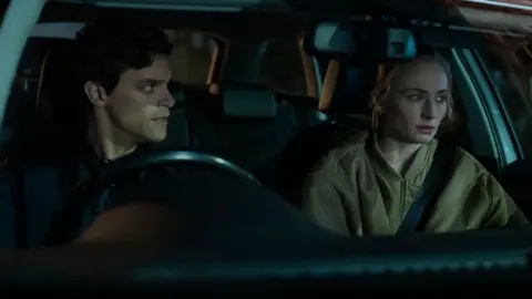 Amazon Content Services LLC Jacob Fortune-Lloyd and Sophie Turner sit in the front of a car at night. Fortune-Lloyd and Turner both have scared looks on their faces. Fortune-Lloyd's outfit is black whilst Turner wears a khaki green bomber jacket and has her seatbelt on.
