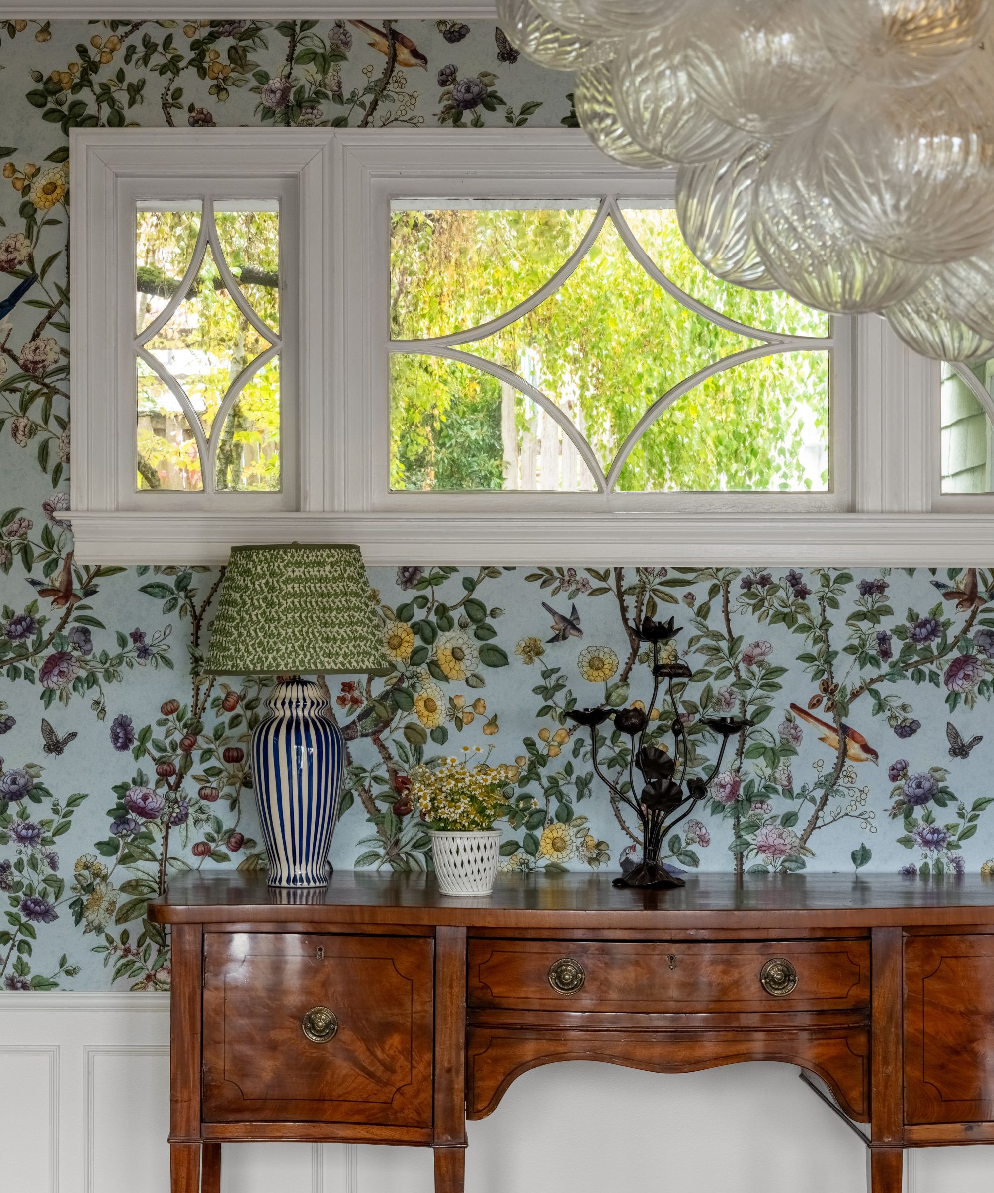 The corner of a room with light blue floral wallpaper, a horizontal window, and an antique wooden console table.