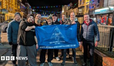 Men in a walking group stand in a shopping street and hold up a banner. It is night-time and there are Christmas lights above the road. Several men are standing near railings and they are wearing jackets, hats and trainers.
