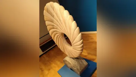 West Northants Council A large stone carving of a circular piece of art is sat on a plinth in a gallery.
It has fluted edges and patterns and there is a hole in the centre of the work.