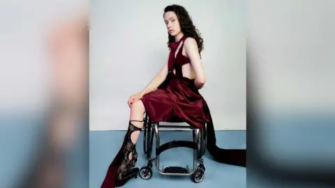 David PH Hyde Alice Dyer has curly brown hair and is sitting sideways in her wheelchair. She's posing for a photo shoot wearing a dark red short dress with cutouts. She has on black lace socks