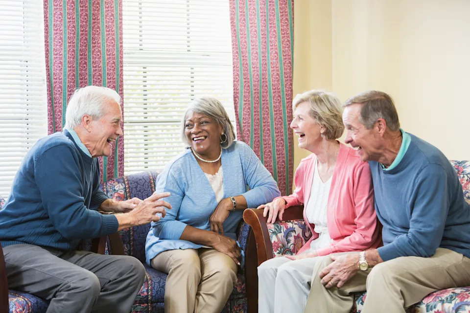 A multi-ethnic group of four seniors sitting together in a living room or cummunity center having a conversation and laughing. A senior couple sitting together on a couch and an African American woman are looking at and listening to their friend talk.