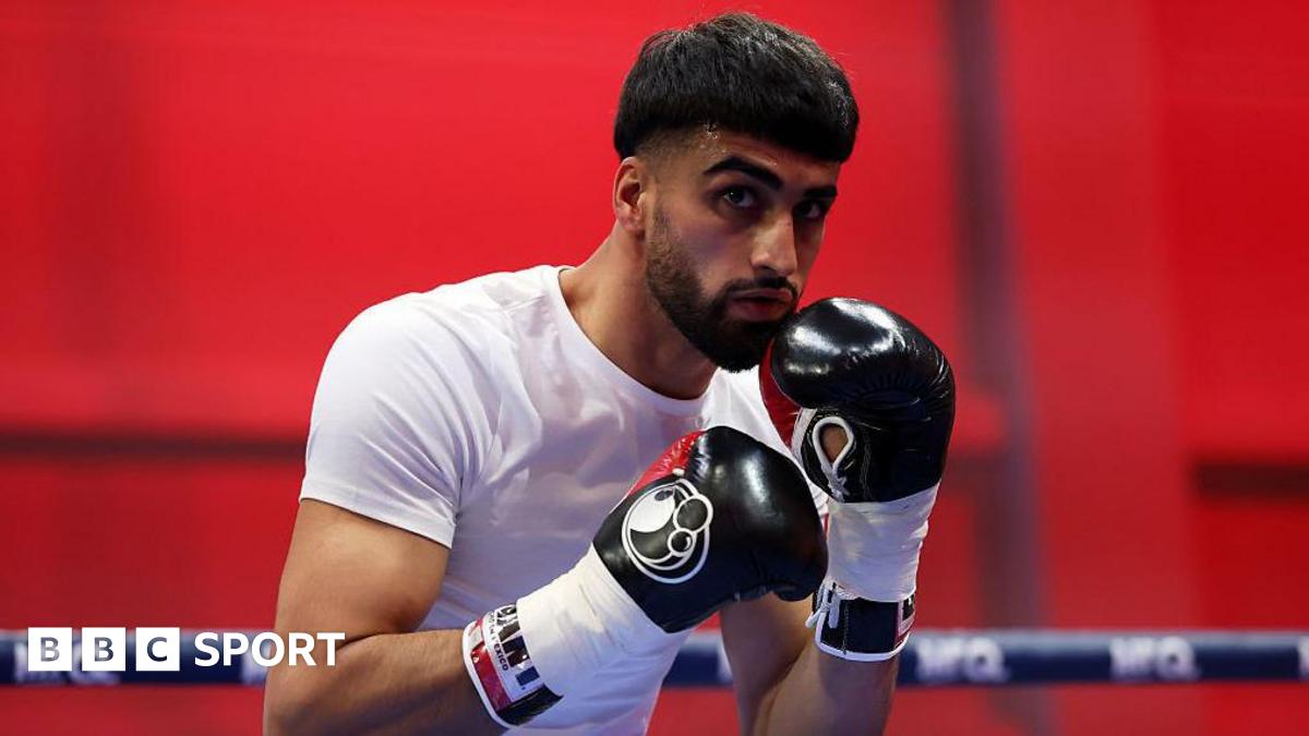 Adam Azim holds up his hands and is wearing a white t-shirt with black boxing gloves