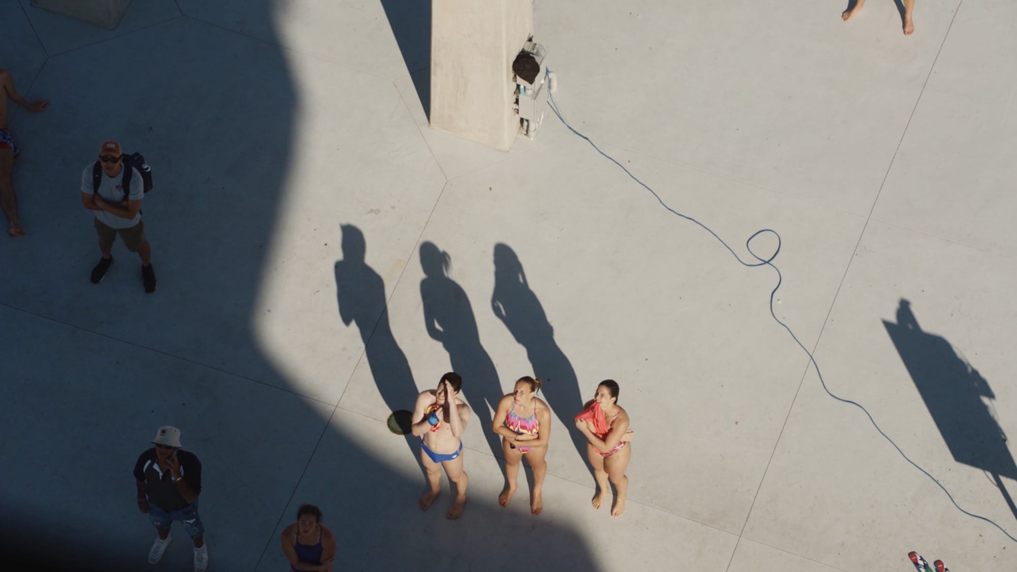 A still from a short film showing three elite women divers standing poolside, viewed from above