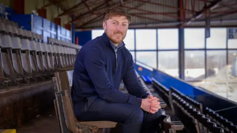 Skills Development Scotland A man with ginger hair and a beard sits on one of the rows of wooden chairs in an old football stand. He is wearing a blue zip up top and blue trousers and has his hands clasped in front of him.