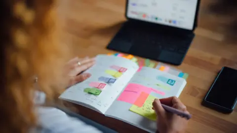 Getty Images A person is planning and writing notes in a journal at a cozy cafe. A laptop and smartphone are on the desk.