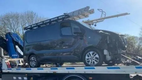 A grey Nissan van, with severe damage to its front, loaded on to a tow truck