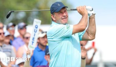 Sir Nick Faldo wearing a light blue t-shirt and a dark blue baseball cap holds a golf club over his shoulder
