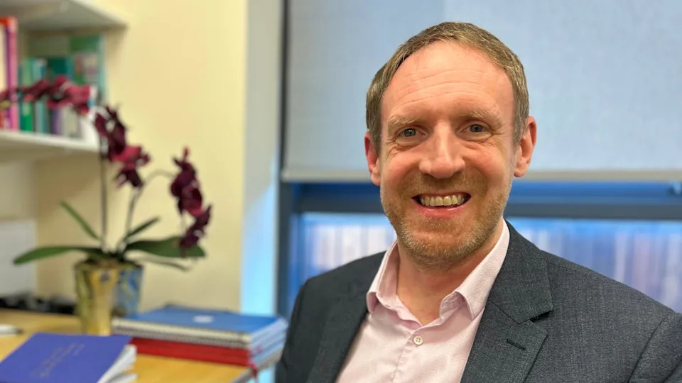 Prof John-Paul Taylor is seated in his office at Newcastle University's Centre for Ageing and Vitality. He is wearing a grey suit and pink shirt. He has brown hair and stubble. Behind him are different coloured exercise books on his desk and books on a shelf.