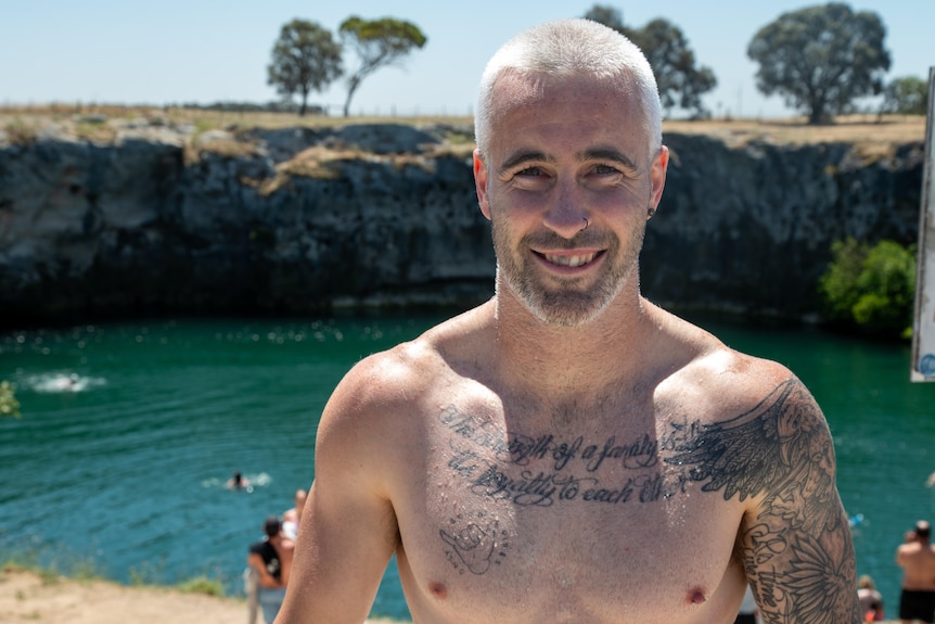 A man without a shirt smiling, with people swimming in a lake behind him