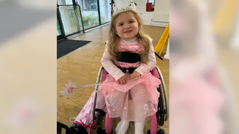 Charlie Brown Dani-Rae Brown is wearing a tiara and a pink princess costume, and holding a wand, while sitting in a wheelchair in a room indoors.
