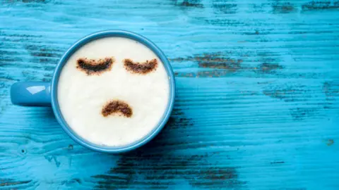 Getty Images A birds-eye view of a blue coffee mug containing a cappuccino. The frothy top is dusted with chocolate in a sad emoji design. The mug sits atop a wooden table painted in a similar cool blue colour.