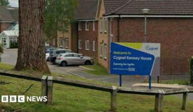 A sign for 'Cygnet Kenney House' in blue and yellow, placed on a small lawn by the entrance to a mental health unit. Cars can be seen parked in a car park while a large tree can be seen on the grass bank.