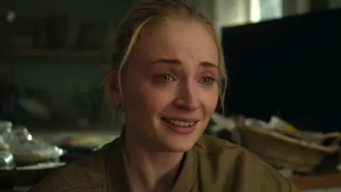 Amazon Content Services LLC Picture of Sophie Turner looking upset, with tears in her eyes and her hair pulled back. She is wearing a khaki bomber jacket that is slightly unzipped. Messy blurred household items appear in the background.