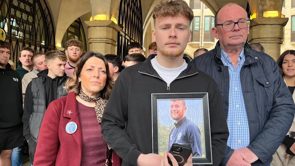 Luke Abrahams' parents Julie Needham and Richard Abrahams are pictured outside the inquest with their younger son, Jake. She has shoulder-length brown hair and is wearing a pink top and red jacket. Richard is on the right and is wearing spectacles, a blue checked shirt and blue jacket. Jake is in the centre, and has short brown hair and a ginger goatee beard. He is wearing a black top and is holding a photo of his brother Luke as well as a mobile phone.