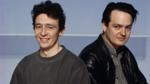 A very young Paul Whitehouse and Charlie Higson - Whitehouse, with a fine head of dark brown hair, wears a grey sweatshirt while Higson, with very dark hair, slicked back, sits in a grey shirt, jeans and a leather jacket. 