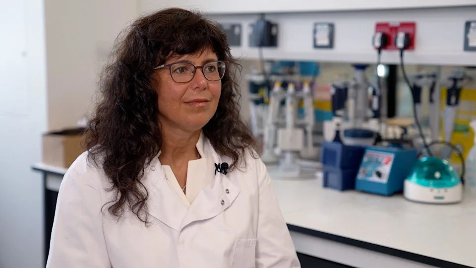 A woman with dark curly hair and glasses is wearing a white lab coat. She is standing in front of a table which is lined with medical an scientific equipment in a clinical lab setting.