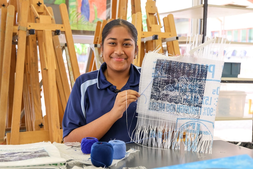A young girl in a blue school uniform smiles while she holds up a print of fish on a phosphate bag and sits in front of easels.