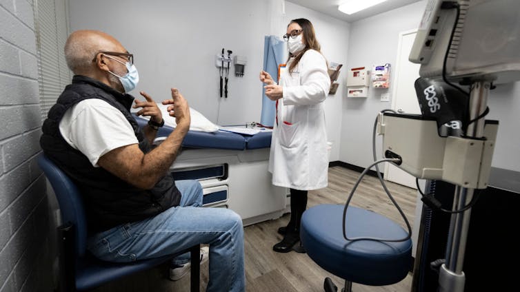 Patient wearing surgical mask sitting in chair of exam room, talking to a doctor