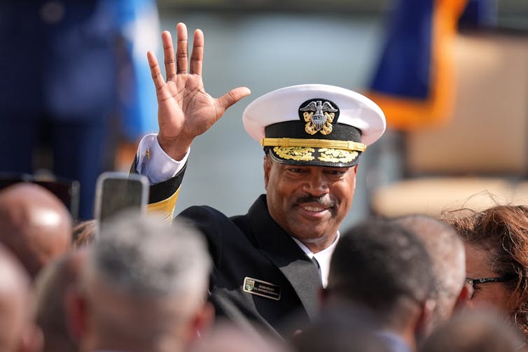 A Black man in a Navy uniform waves and smiles.