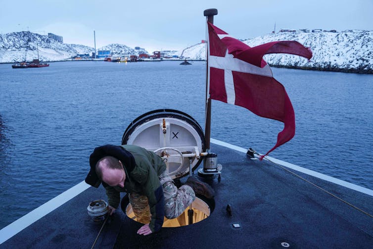 A man climbs out of a hatch on the bow of a ship. A red and white flag flies beside him.