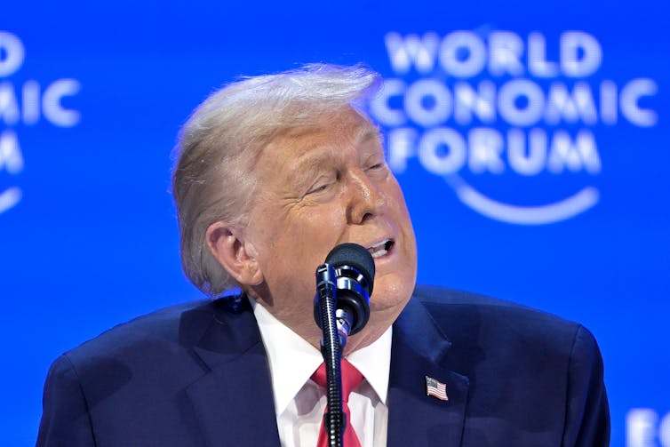 An older man with fluffy white-blond hair contorts his face while speaking into a microphone.