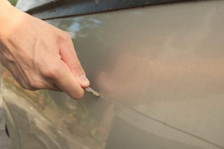 A hand holding a car key traces it along the side of a beige-colored car to leave a scratch.
