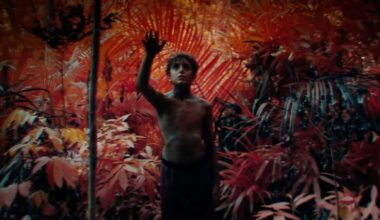 young barechested boy in a forest lit with exotic orange and red hues, holding up one hand as if yo touch something only he can see