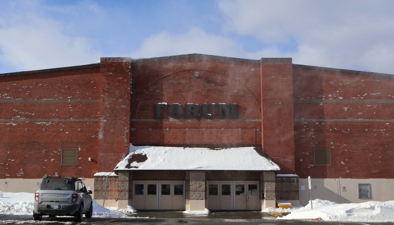 A red brick building with a sign that says "Forum" above the main entrance. That entrance has several beige doors and a canopy over it covered in a dusting of snow. An SUV sits in the parking lot to the left.