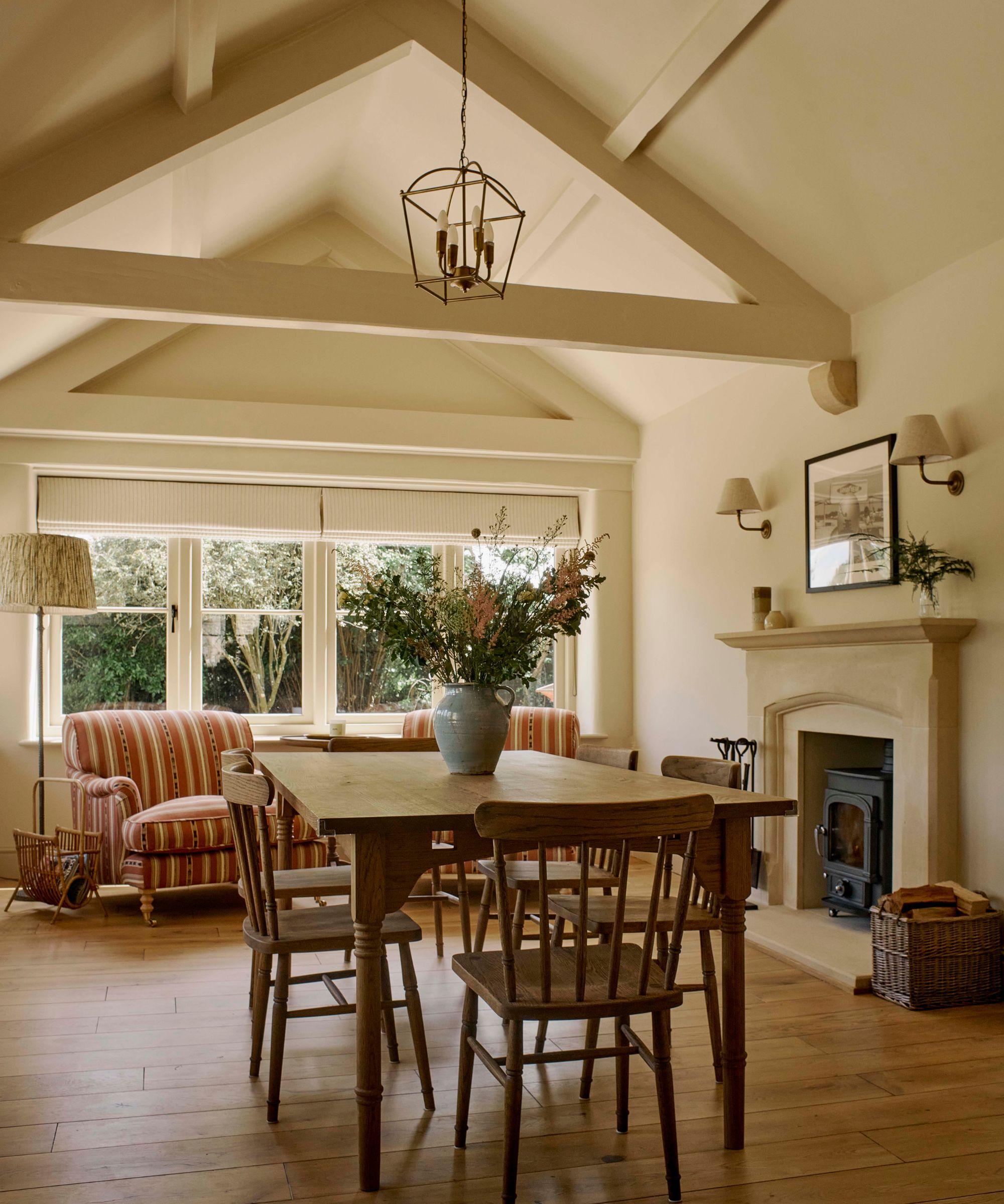 A warm neutral dining room with tall pitched ceilings, a wooden table and chairs, and two striped armchairs by the window
