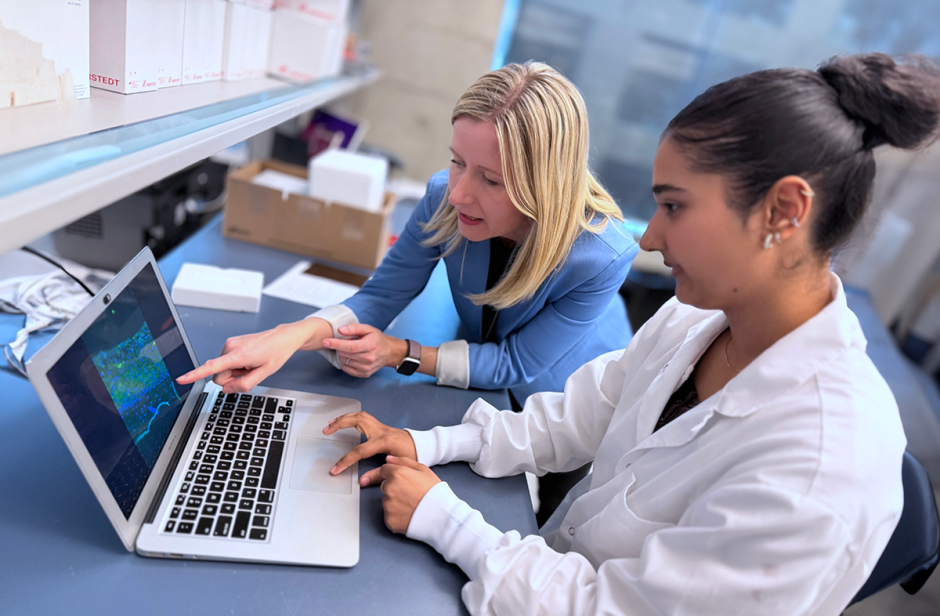 Two individuals in a lab setting look at a laptop. One points at the screen. 