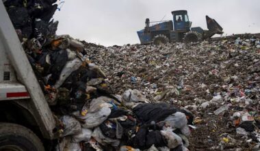 A compactor drives over a large pile of mixed trash and plastic bags at a landfill site, with waste spilling from a garbage truck in the foreground.