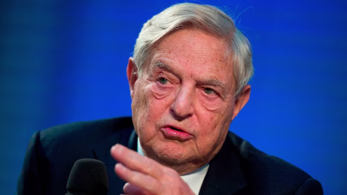 George Soros speaks while gesturing with his hand against a blue background.