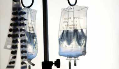 Two intravenous fluid bags hang from a metal stand with medication packaging strips nearby.