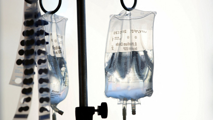 Two intravenous fluid bags hang from a metal stand with medication packaging strips nearby.
