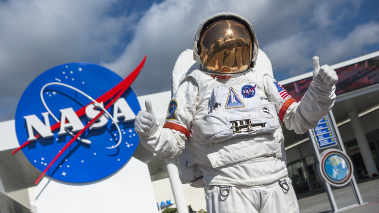 An astronaut suit flashing two thumbs up in front of the Kennedy Space Center