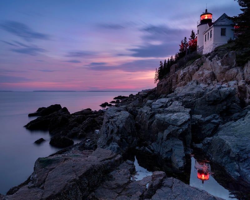 A lighthouse on a rocky cliff overlooks calm water at sunset, with pink and purple hues in the sky and reflections in tide pools below. Pine trees surround the lighthouse, and its red light is illuminated.