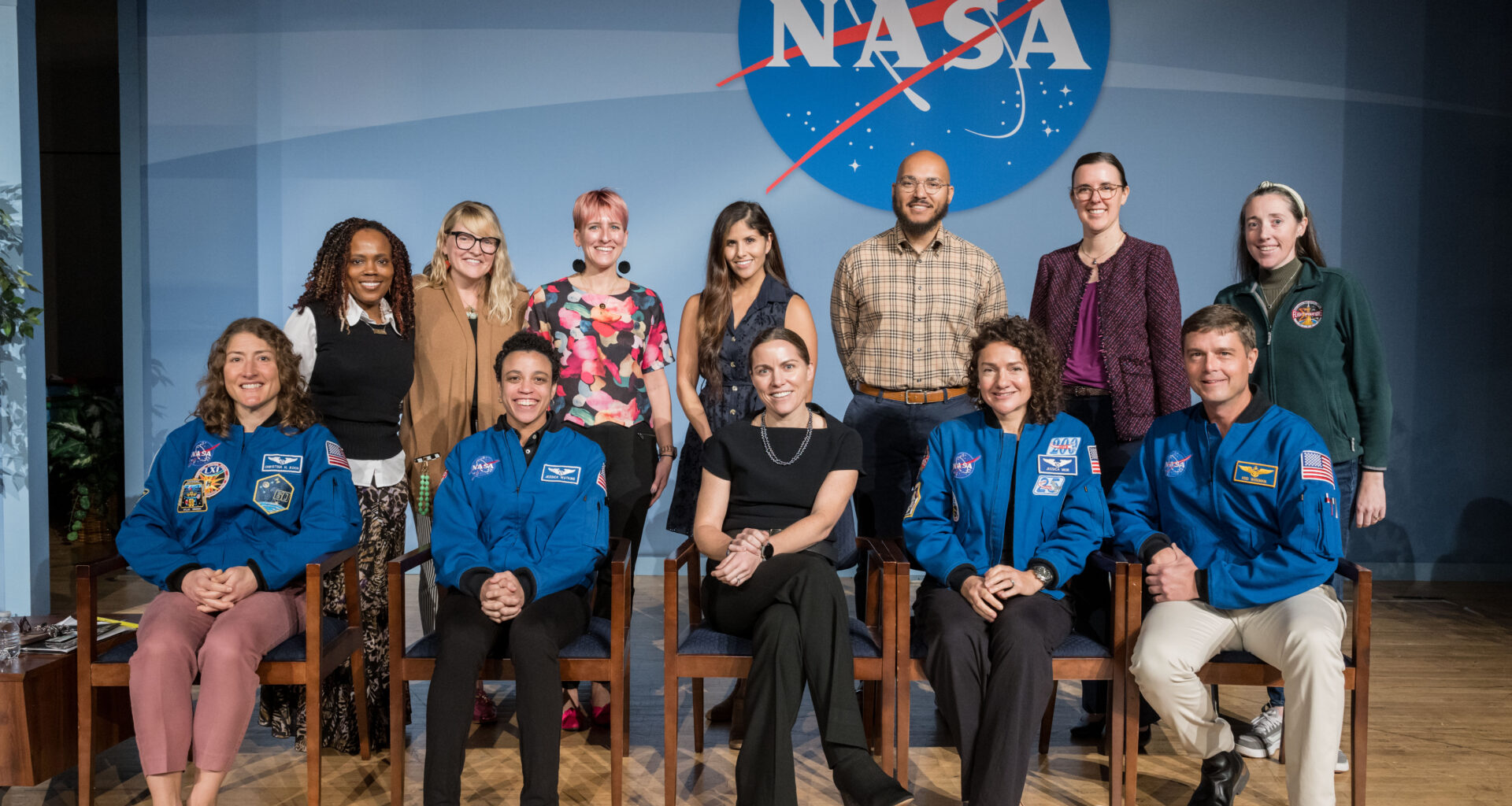 A group of people pose in an auditorium in front of a blue background with a NASA meatball insignia.