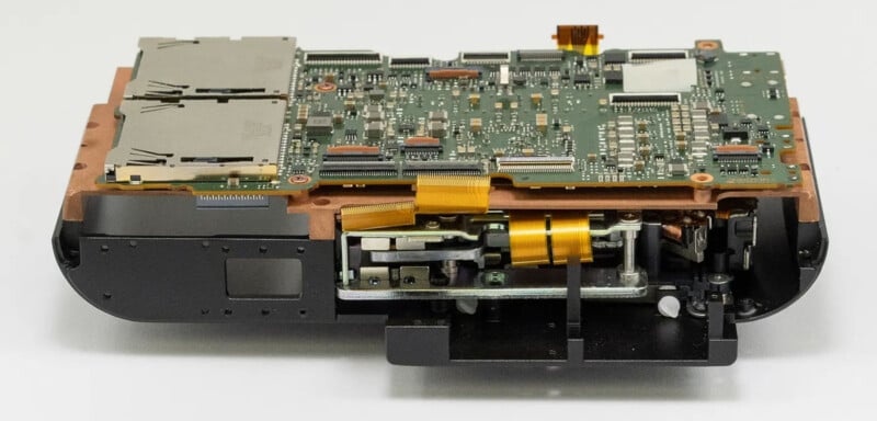 Close-up of the internal components of a digital camera, showing a circuit board, ribbon cables, and metal mechanical parts on a white background.