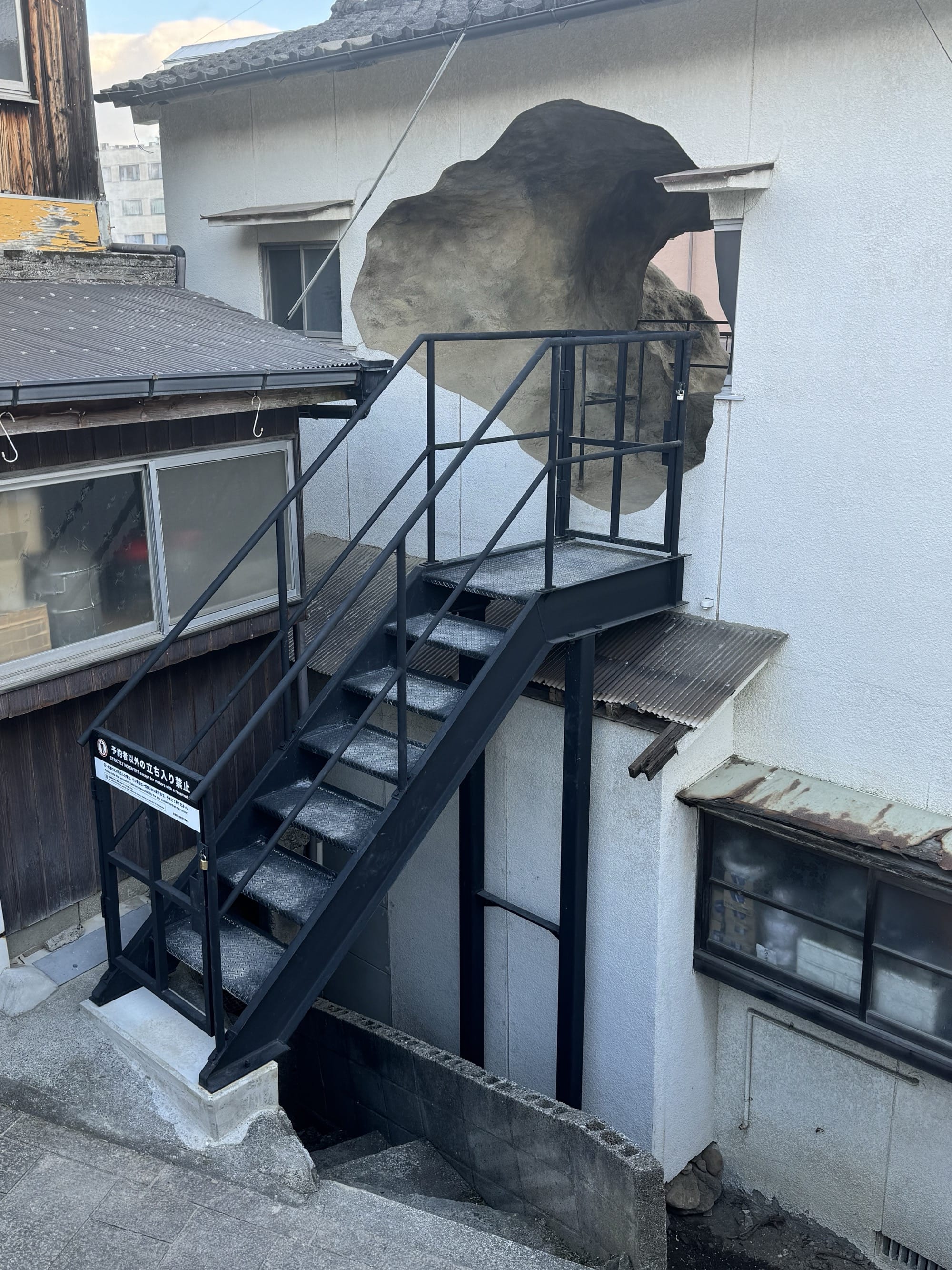 A detail of an art installation comprising a black staircase leading into an upper level of a small house with holes in it that open to what looks like a cave on the inside
