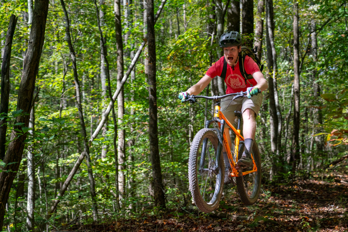 A mountain biker jumps over a small section of a dirt path in a lush green forest. The cyclist, wearing a helmet and a red t-shirt, shows an expression of excitement as he rides an orange mountain bike. Surrounding trees and foliage create a vibrant and natural backdrop.