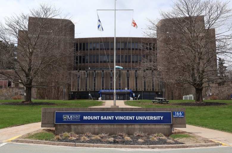 A blue sign with white font that says "Mount Saint Vincent University" hangs from a short stone wall at the base of a small hill with a concrete path running down the centre. In the background is a multi-storey concrete building. A blue sign over the main entrance says "Seton Academic Centre." There are leafless trees and a flag pole on a concrete pad in the grassy centre.