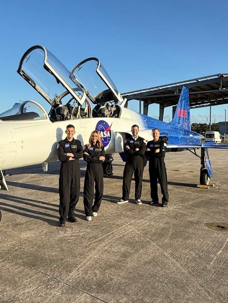 Four people in flight suits stand in front of a white and blue NASA jet with open cockpits.