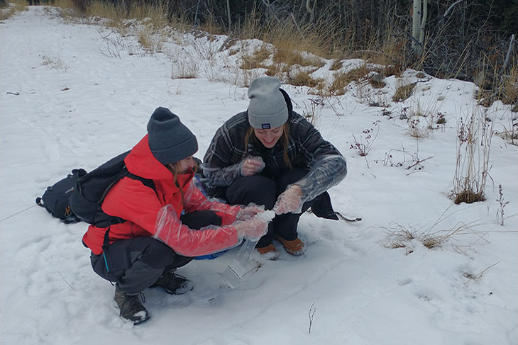 Two people crouching on snow-covered ground in an outdoor setting with dry grass and trees in the background.