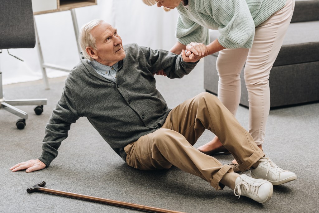 An elderly woman helps her husband, who has fallen onto the floor, get back up.