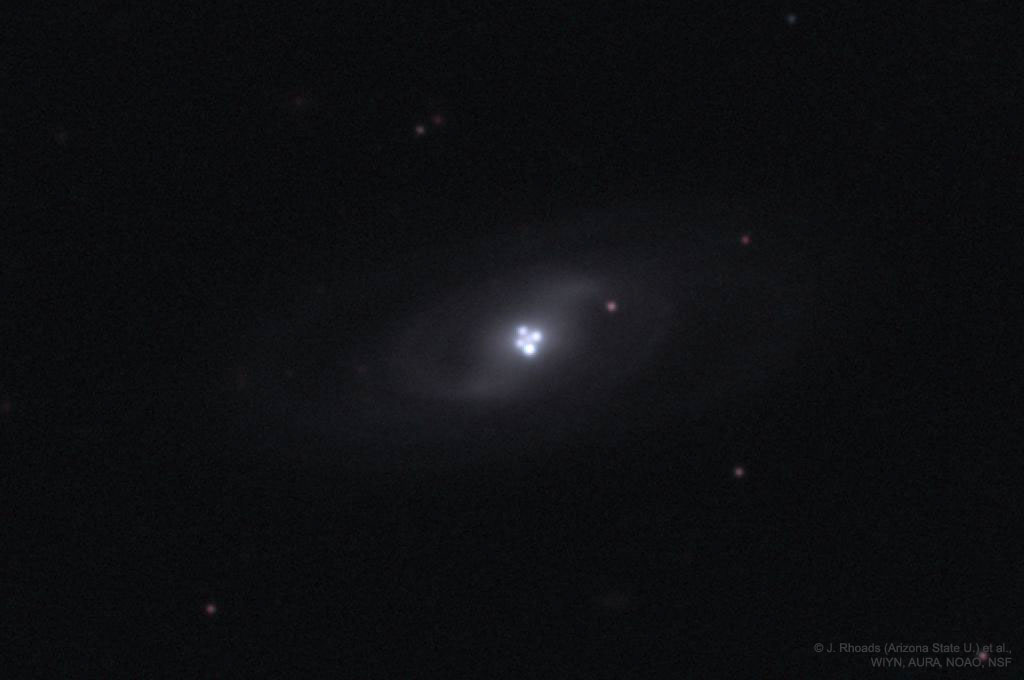 A spiral galaxy with four bright points of light at its center, forming a gravitational lens cross, is surrounded by faint stars against a dark background.